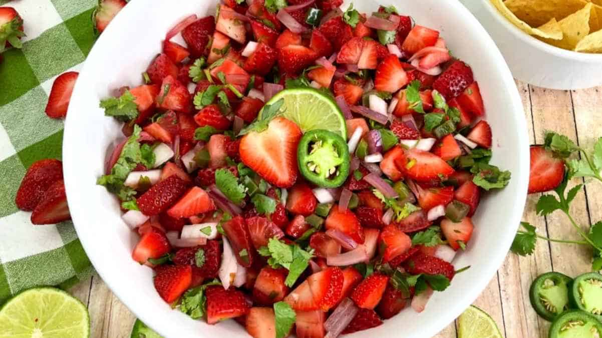 A bowl of strawberry salsa with diced onions, cilantro, lime wedges, and jalapeño slices, served on a wooden table with a green cloth and tortilla chips in the background.
