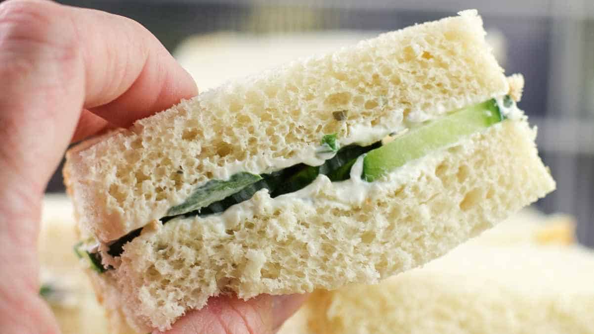 A hand holding a cucumber sandwich with a bite taken out of it.