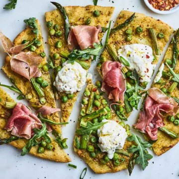 Flatbread pizza topped with arugula, peas, cheese, and prosciutto on a marble surface.