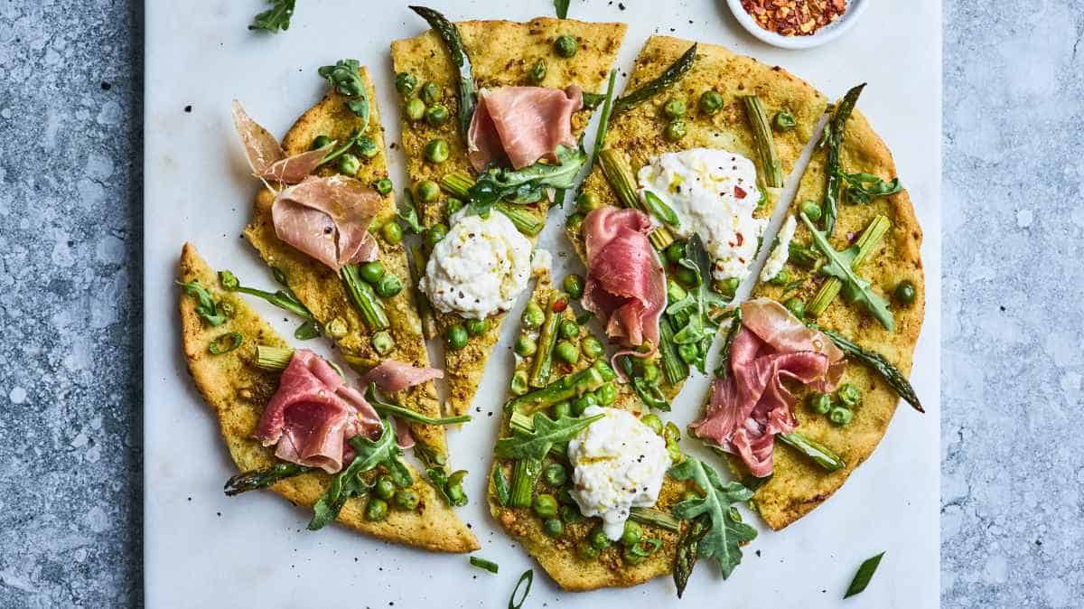 Flatbread pizza topped with arugula, peas, cheese, and prosciutto on a marble surface.
