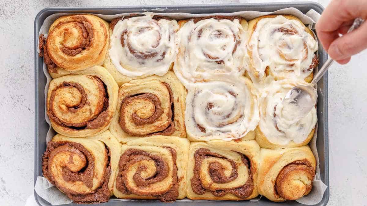 Freshly baked cinnamon rolls with icing being applied to some of them.