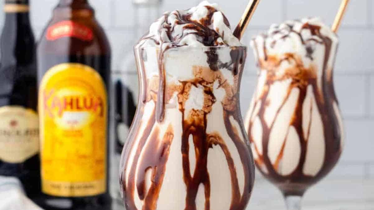 Two glasses of layered dessert topped with whipped cream and chocolate syrup, with bottles in the background.