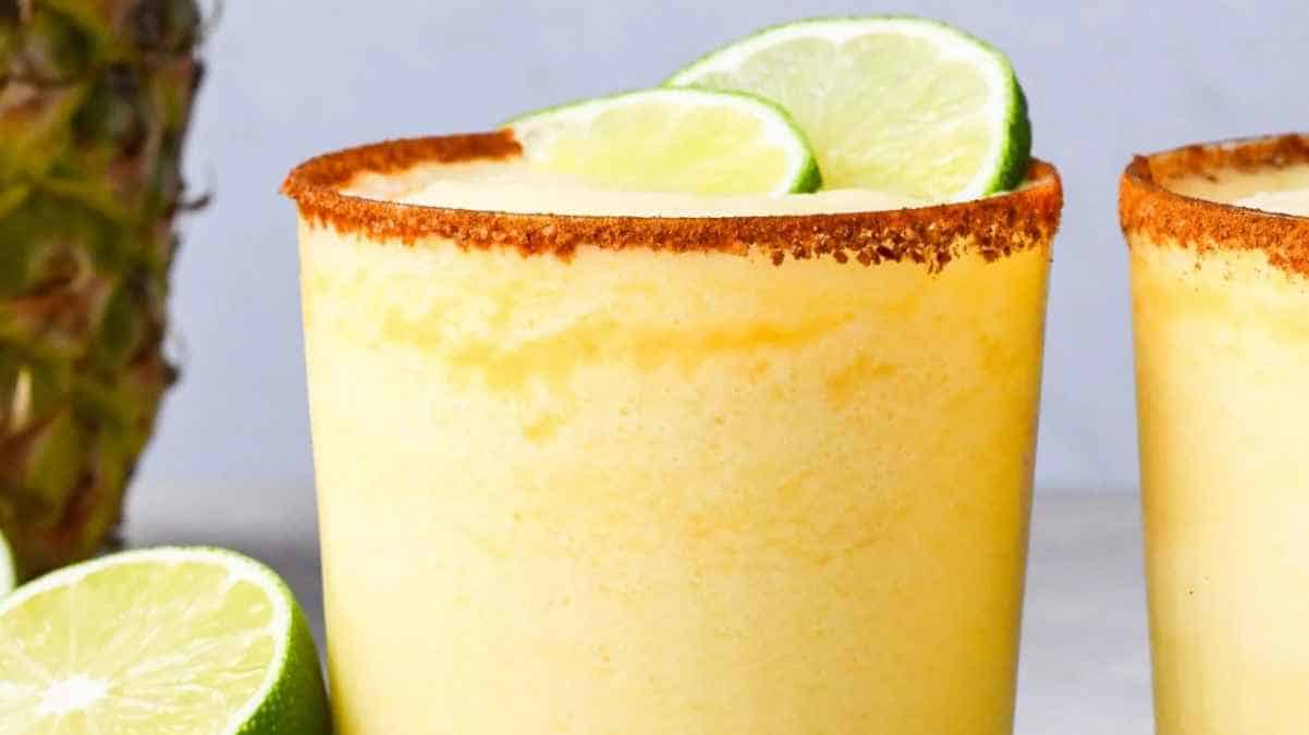 Two glasses of pineapple smoothie garnished with lime slices and a spice-coated rim, with a pineapple in the background.