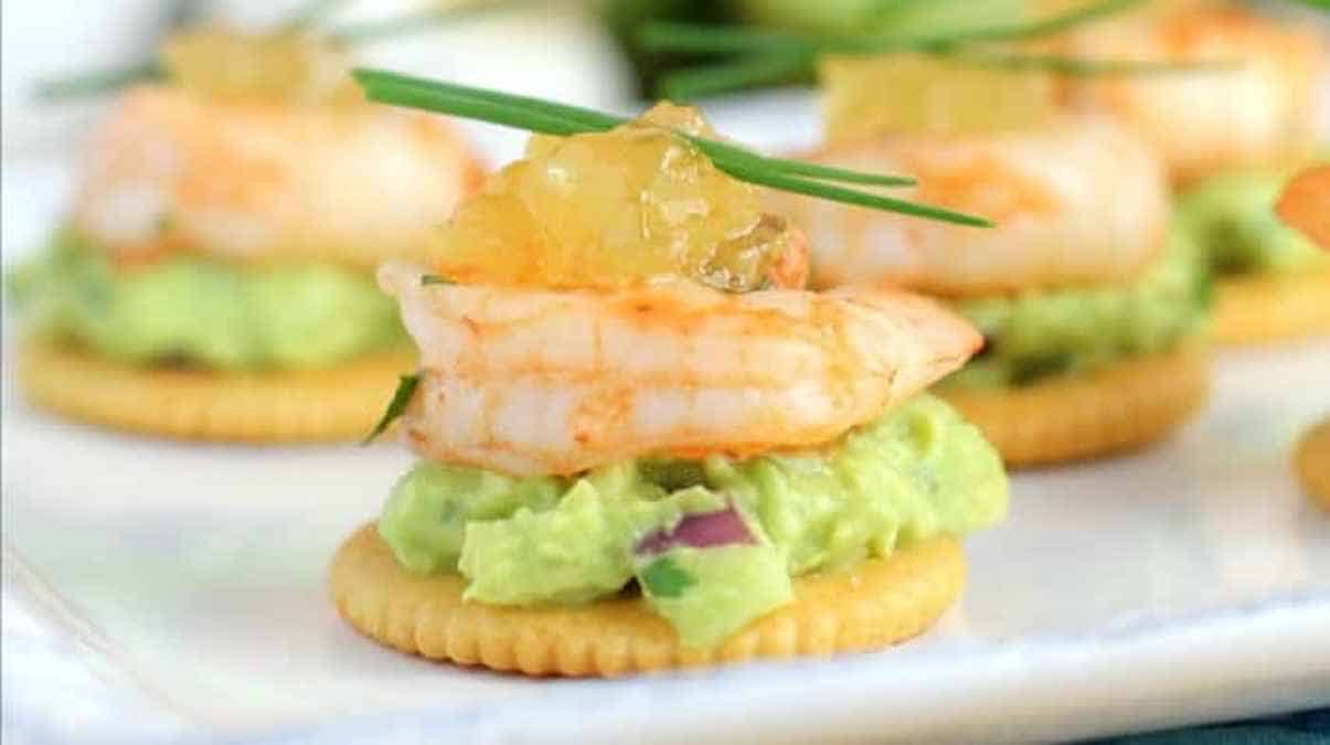 Appetizers with shrimp, guacamole, and a dollop of sauce on crackers, garnished with chives.