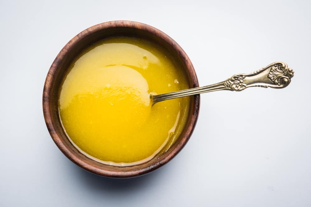 A wooden bowl containing melted ghee and garlic with a metal spoon on a white background.