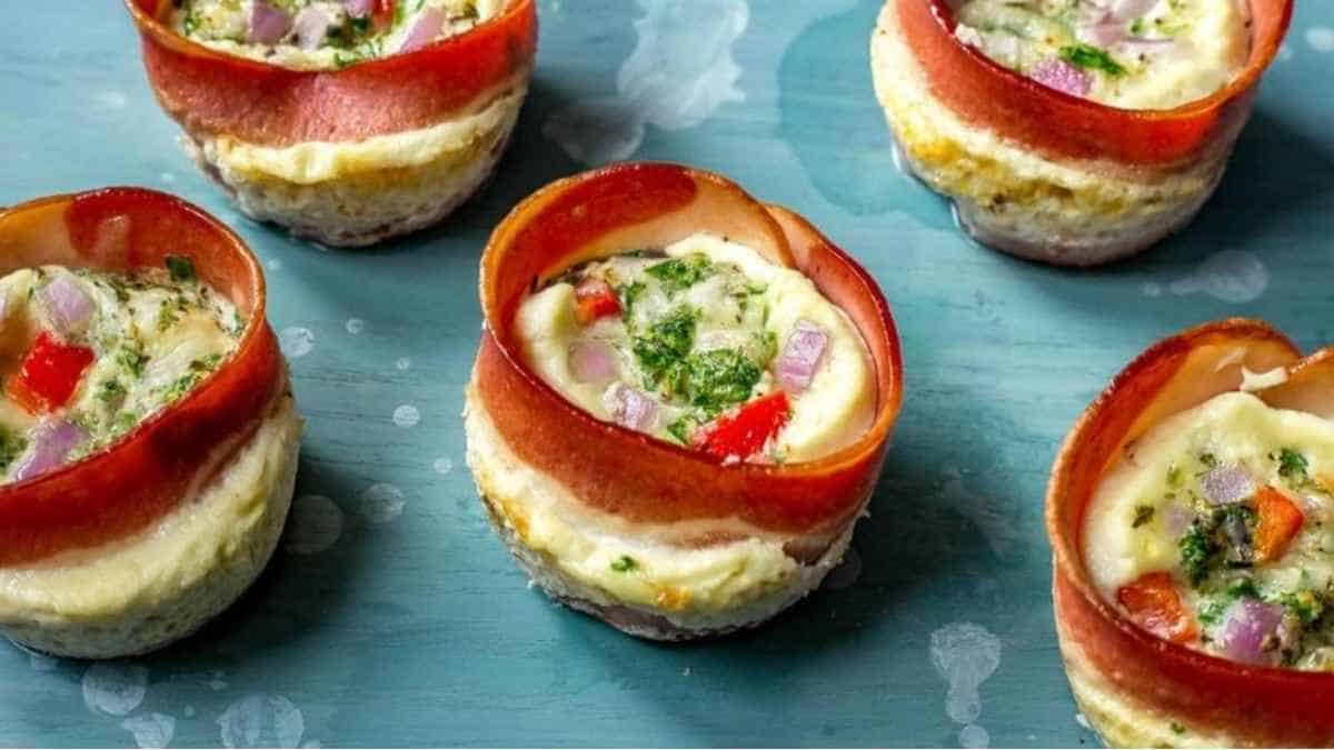 Four egg cups with bacon and vegetables on a blue plate.