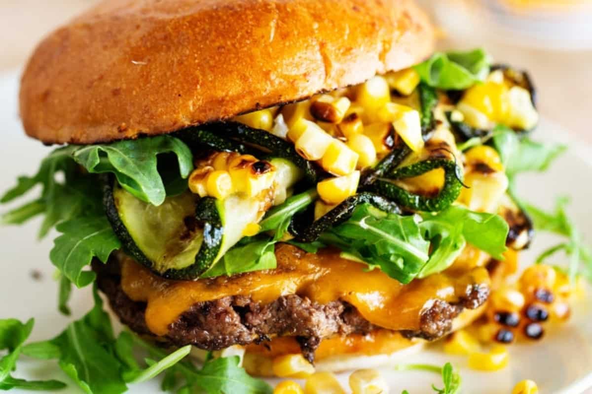 Gourmet cheeseburger with arugula, grilled onions, corn, and sauce on a plate.