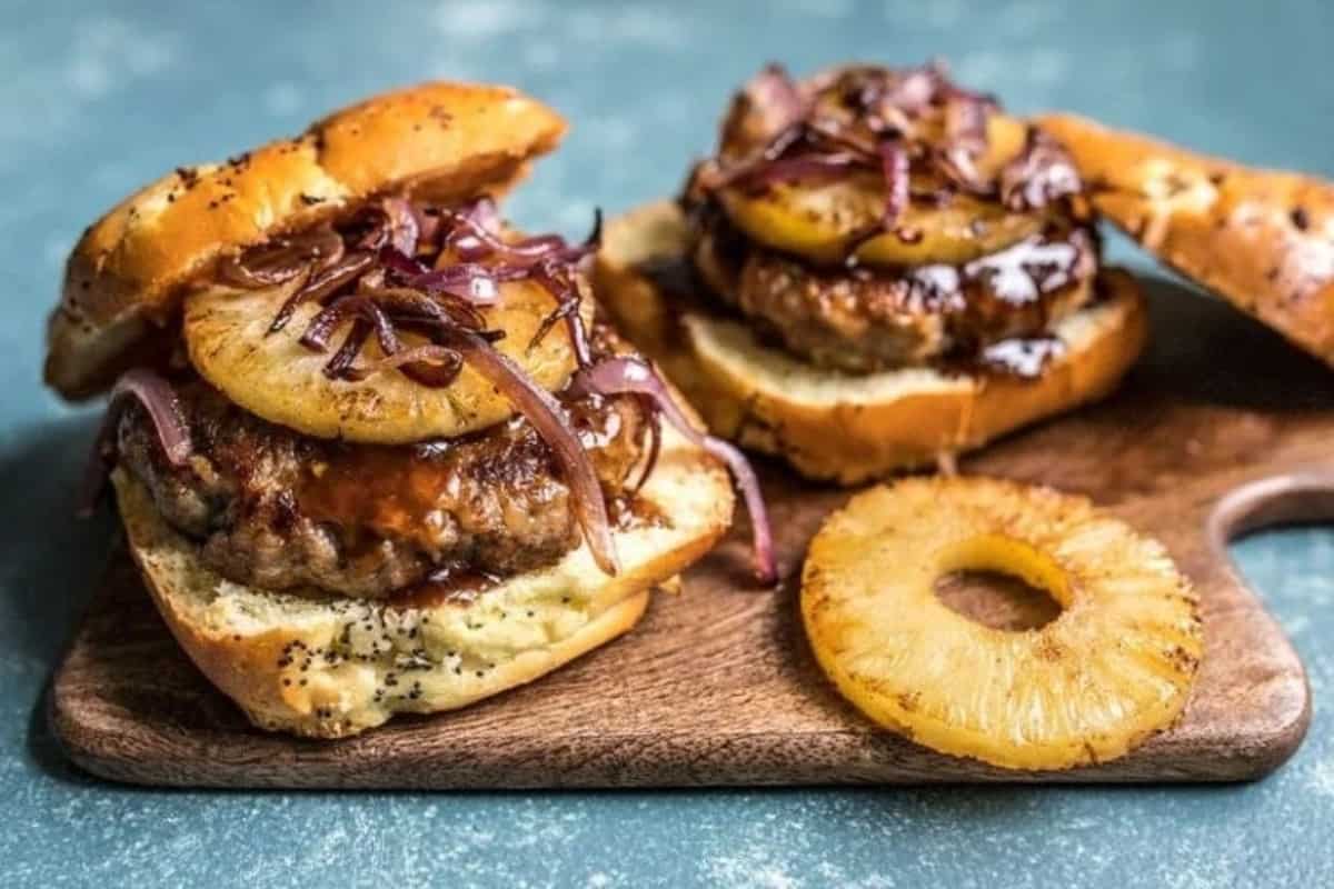 Gourmet burgers topped with grilled pineapple and caramelized onions on a wooden serving board.
