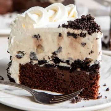 A slice of oreo cheesecake with a chocolate biscuit base and whipped cream on top, served on a white plate with a fork.