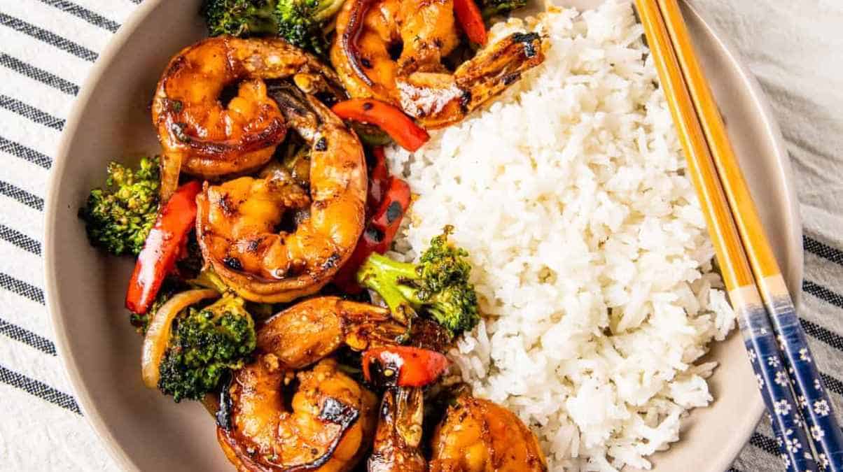 A plate of stir-fried shrimp with broccoli and bell peppers served with white rice and a pair of chopsticks on the side.