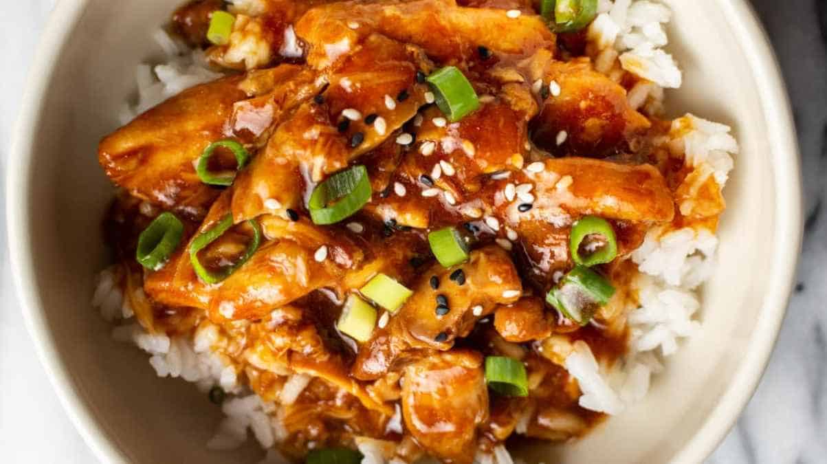 Bowl of rice topped with sesame chicken and garnished with scallions and sesame seeds.