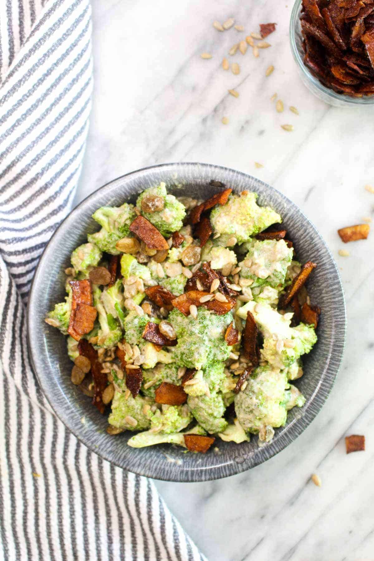 This Healthy Vegan Broccoli Salad is an easy recipe that is BIG on flavor. Broccoli is coated in a sweet and creamy dressing and jazzed up with smokey coconut bacon, toasty seeds, and sweet raisins. 
