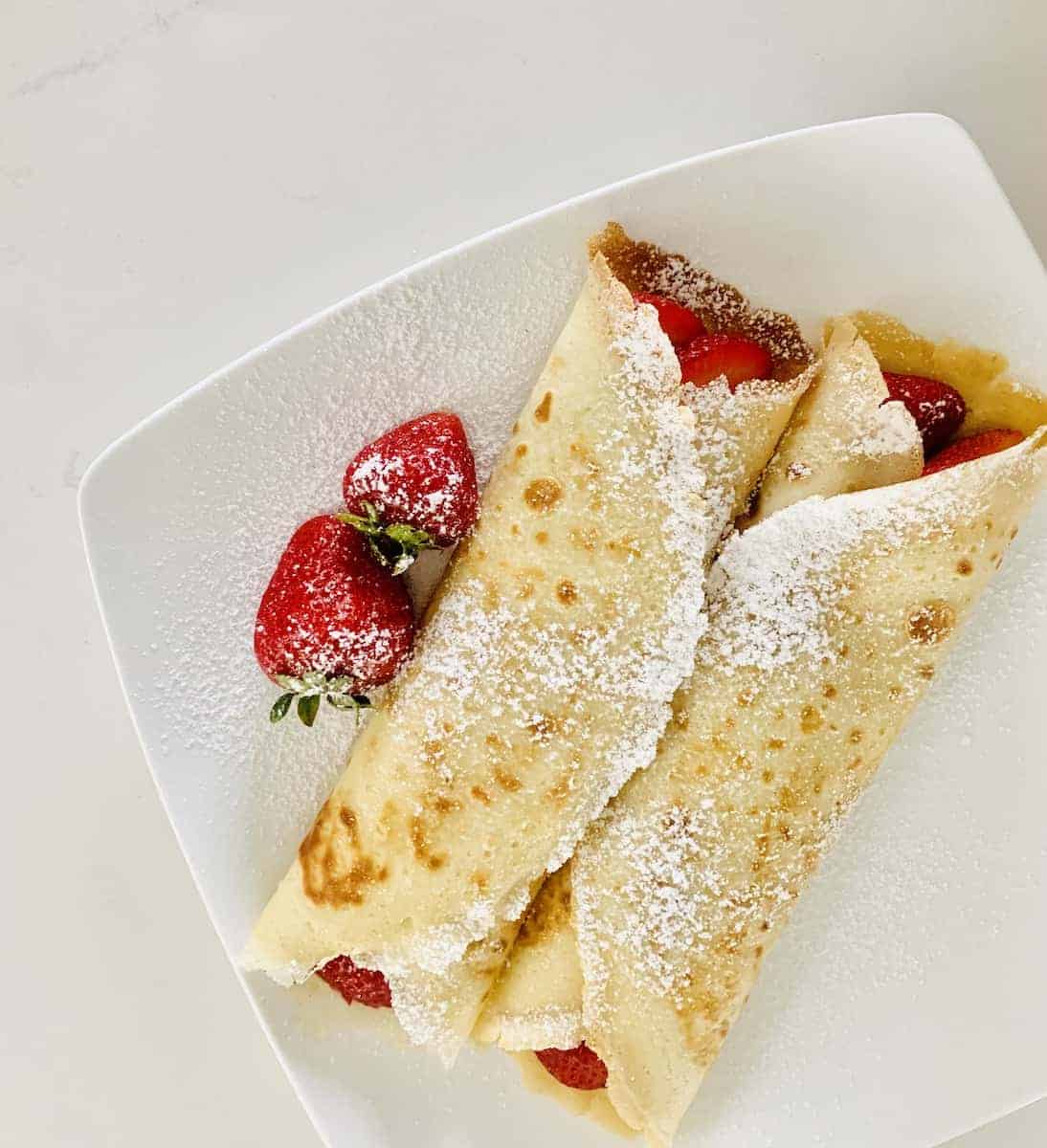 Birds eye view of 2 crepes filled with strawberries sprinkled with powdered sugar, and two strawberries also on the white plate. Red text: "Gluten-Free Crepes". 
