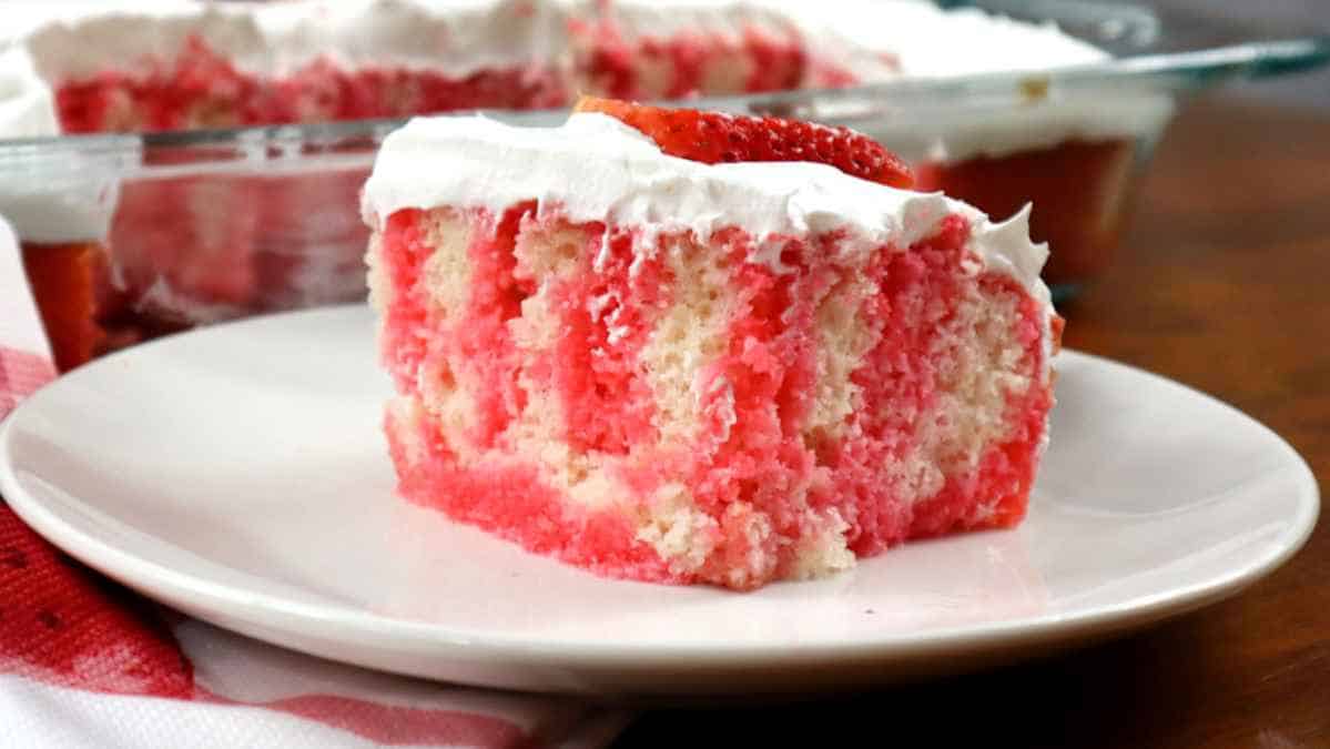 A slice of strawberry poke cake with whipped cream topping on a white plate.
