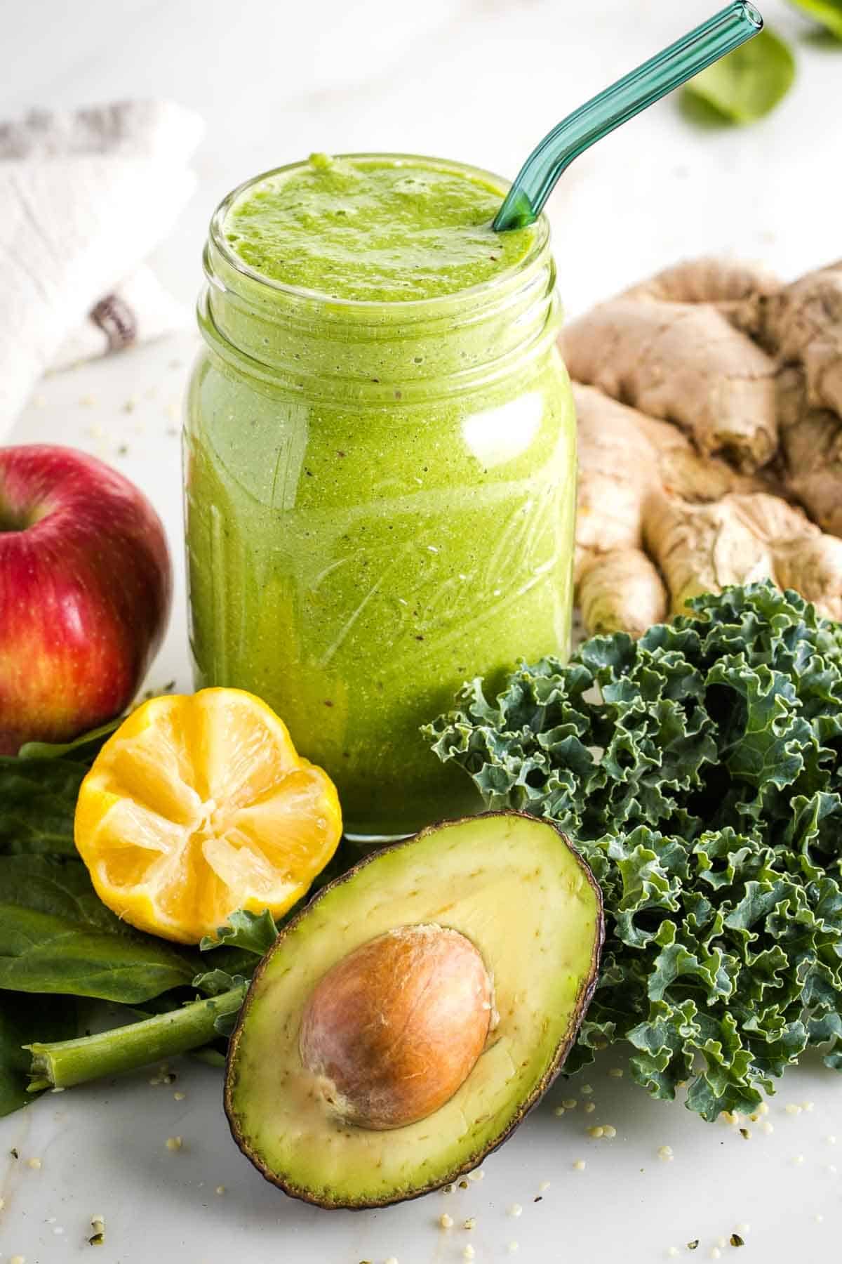 Green smoothie in a mason jar surrounded by greens, lemon, apple, ginger and avocado. 
