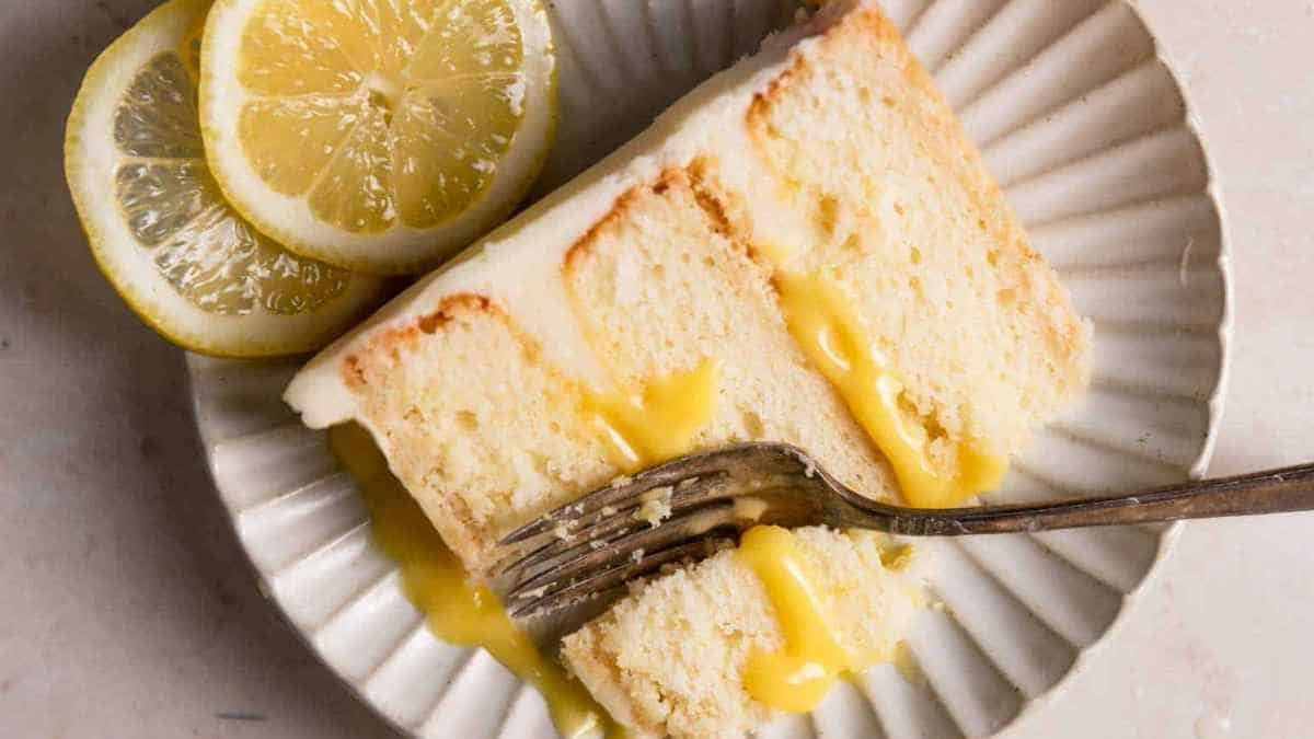A slice of lemon cake with lemon glaze on a plate with a fork and lemon slices.