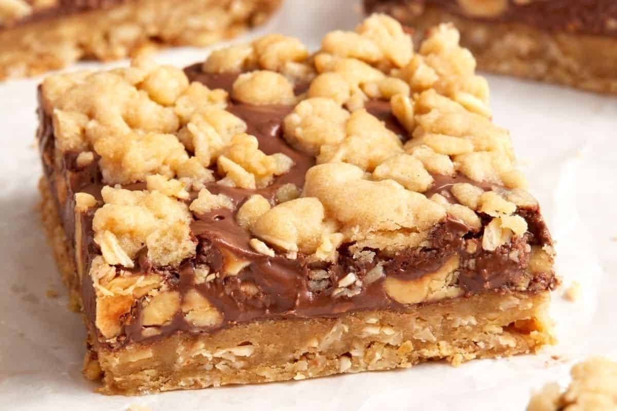 A close-up of a layered dessert bar with a crumbly topping, chocolate layer, and a cookie base.