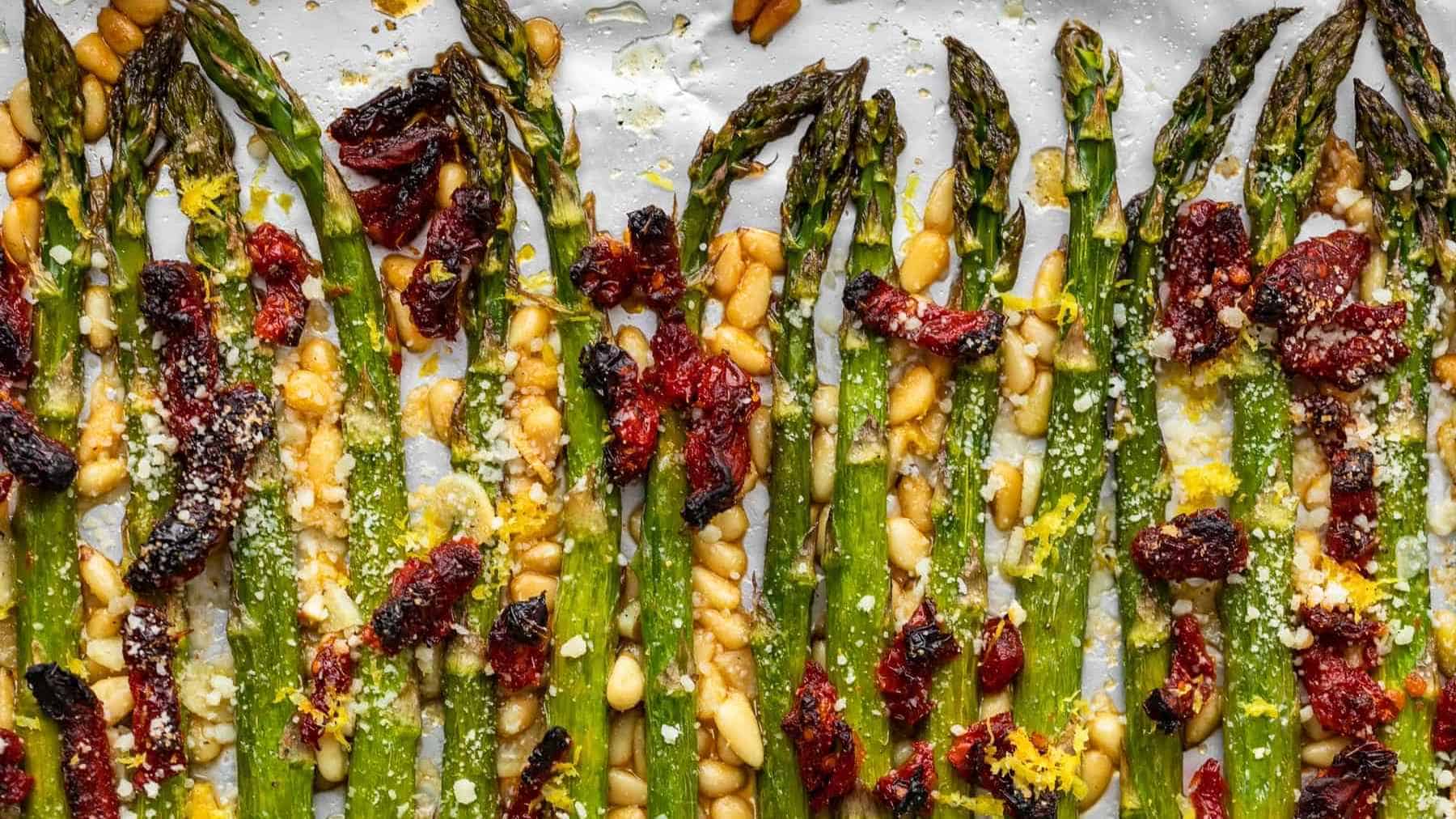 Roasted asparagus with pine nuts, sun-dried tomatoes, and grated parmesan cheese on a baking sheet.