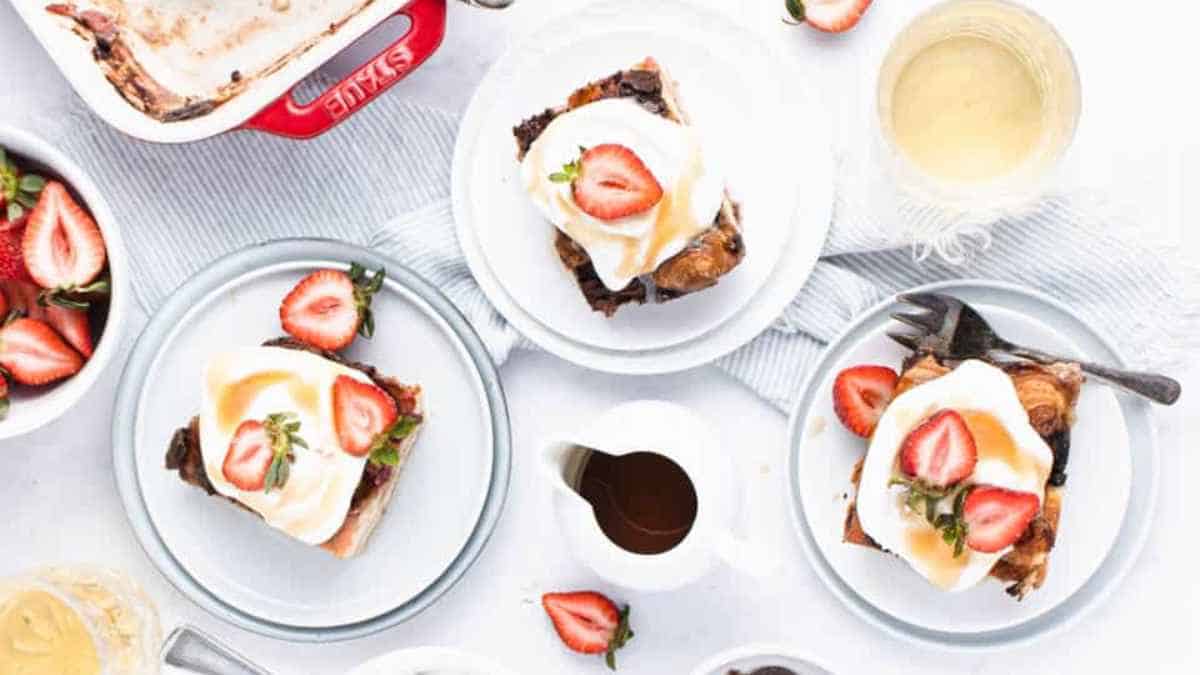 Sliced chocolate cake topped with cream and fresh strawberries served on white plates with extra strawberries and dessert sauce on the side.