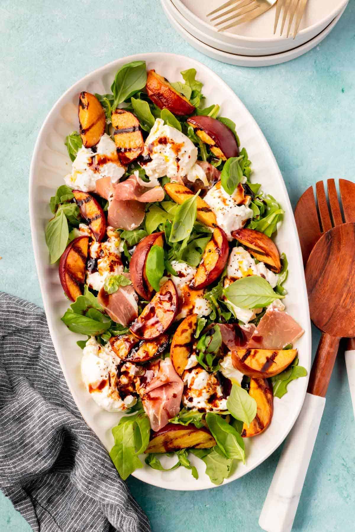 An oval platter filled with grilled peach burrata salad topped with arugula, basil, prosciutto, balsamic glaze, salt and pepper. 
