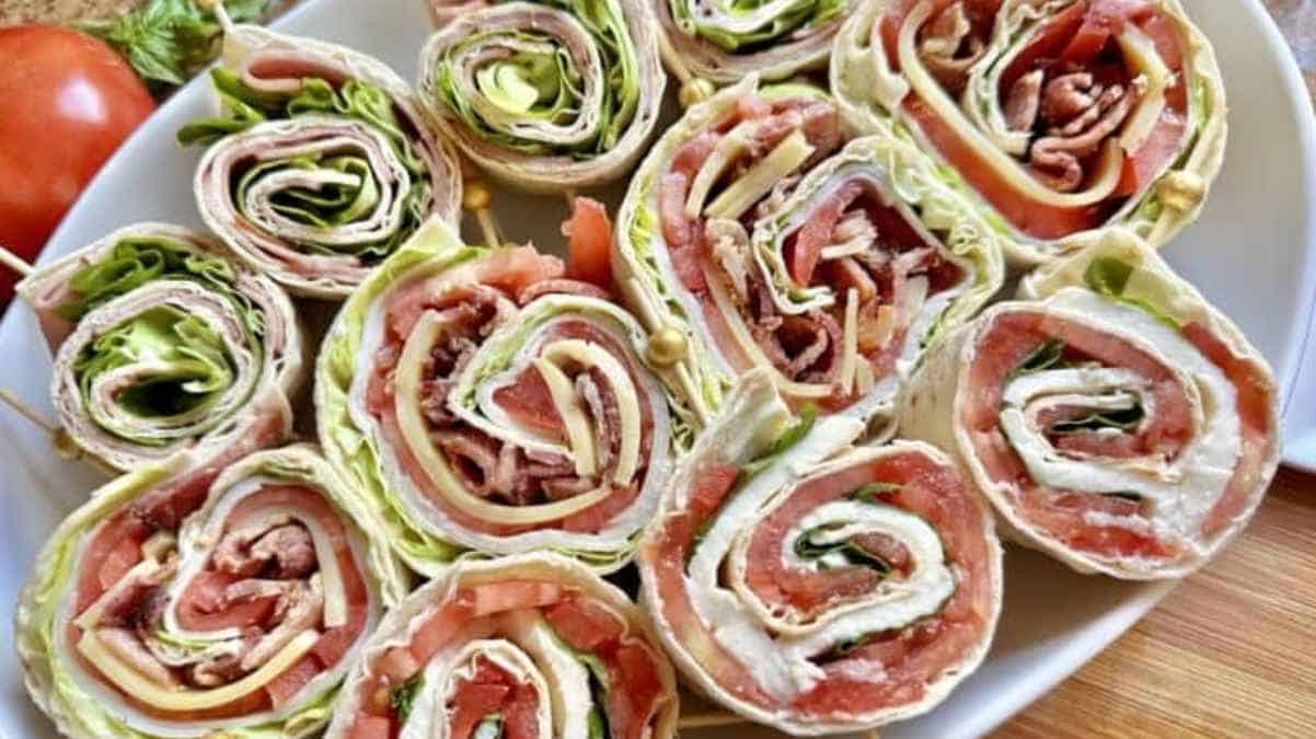 Platter of ham and cheese roll-ups with lettuce, displayed with a whole tomato and garlic cloves in the background.