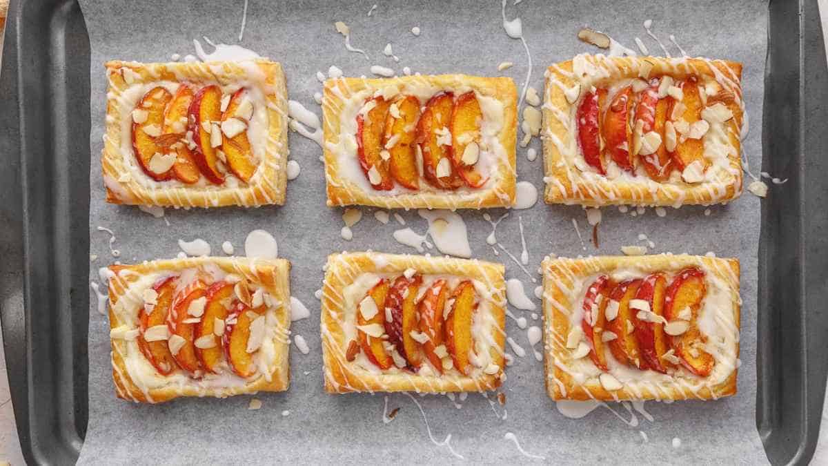 Freshly baked apricot pastries with almond slices and icing drizzle on parchment paper.