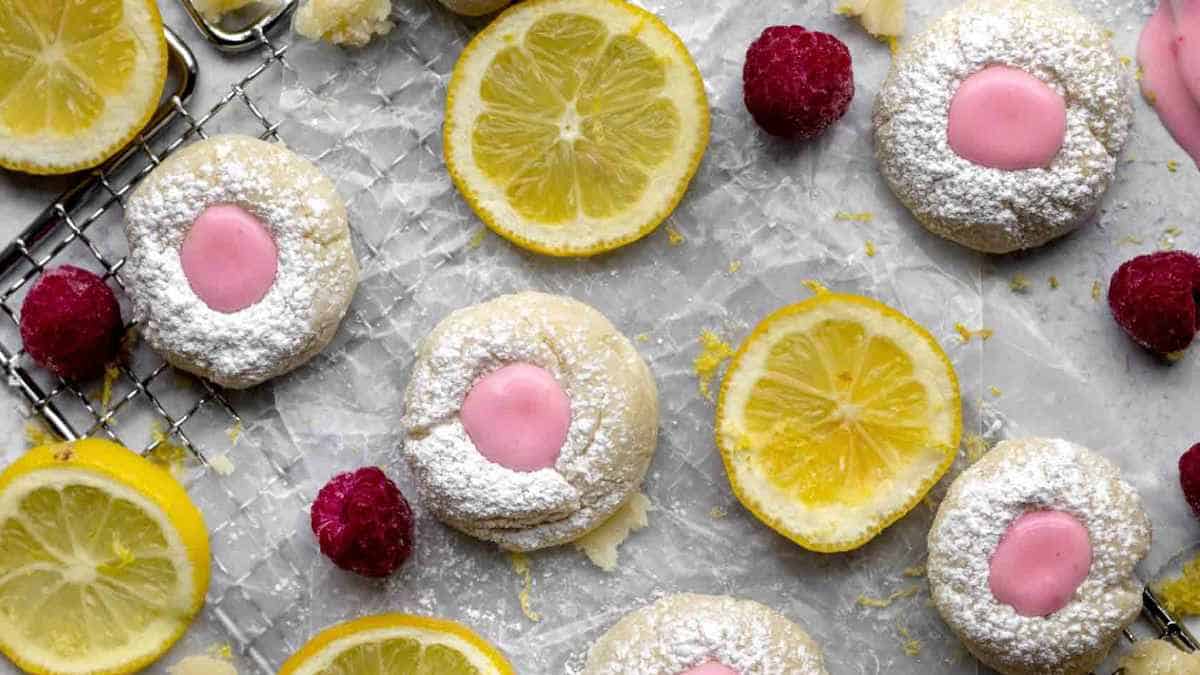 Homemade cookies with pink icing and powdered sugar, garnished with fresh lemon slices and raspberries on a light surface.