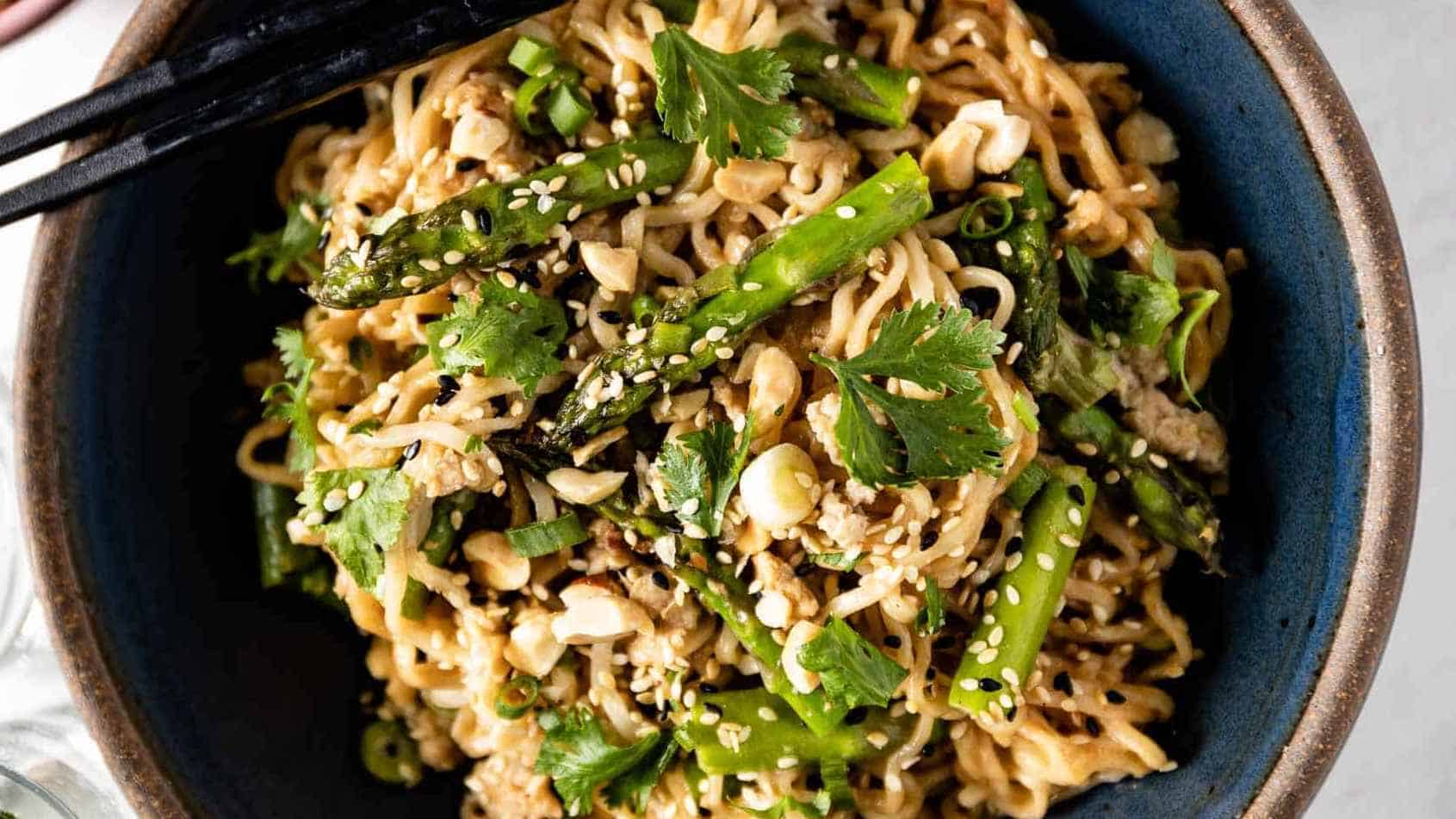 A bowl of stir-fried noodles garnished with green vegetables, sesame seeds, and fresh coriander.