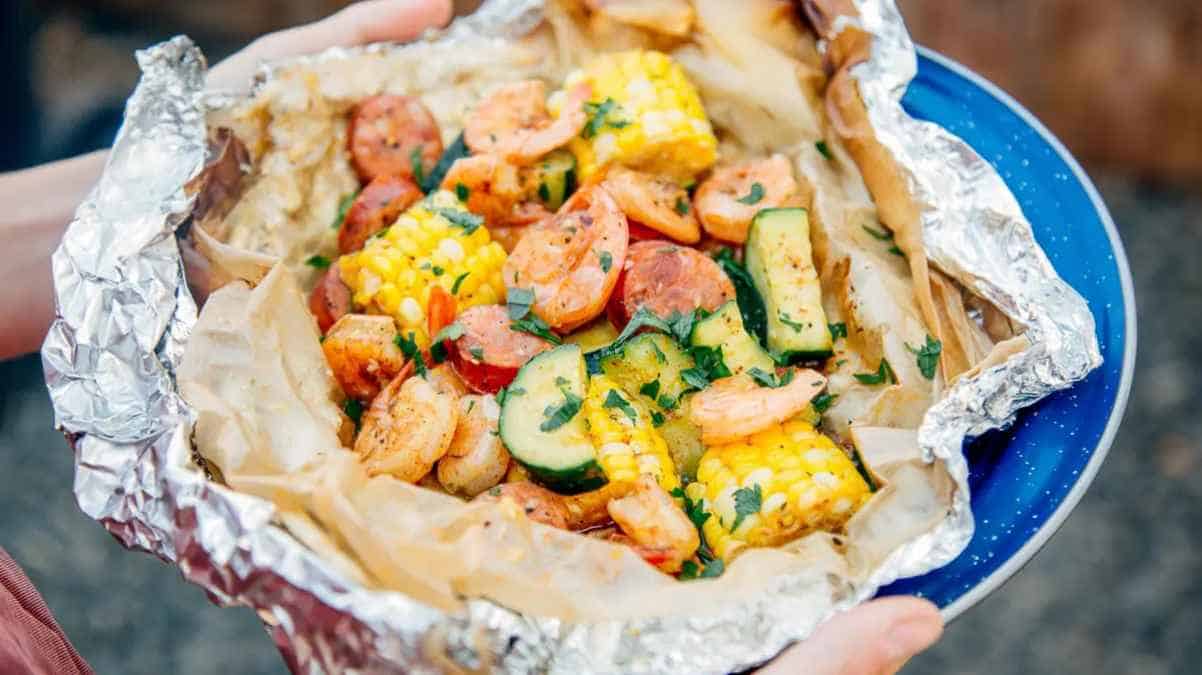 A person holding a plate with a foil packet of shrimp boil containing shrimp, sausage, corn, and zucchini.