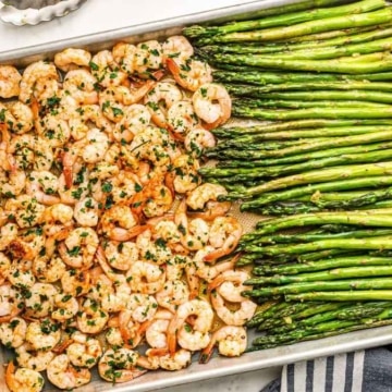 A tray of roasted shrimp and asparagus garnished with herbs, alongside lemon and pasta.