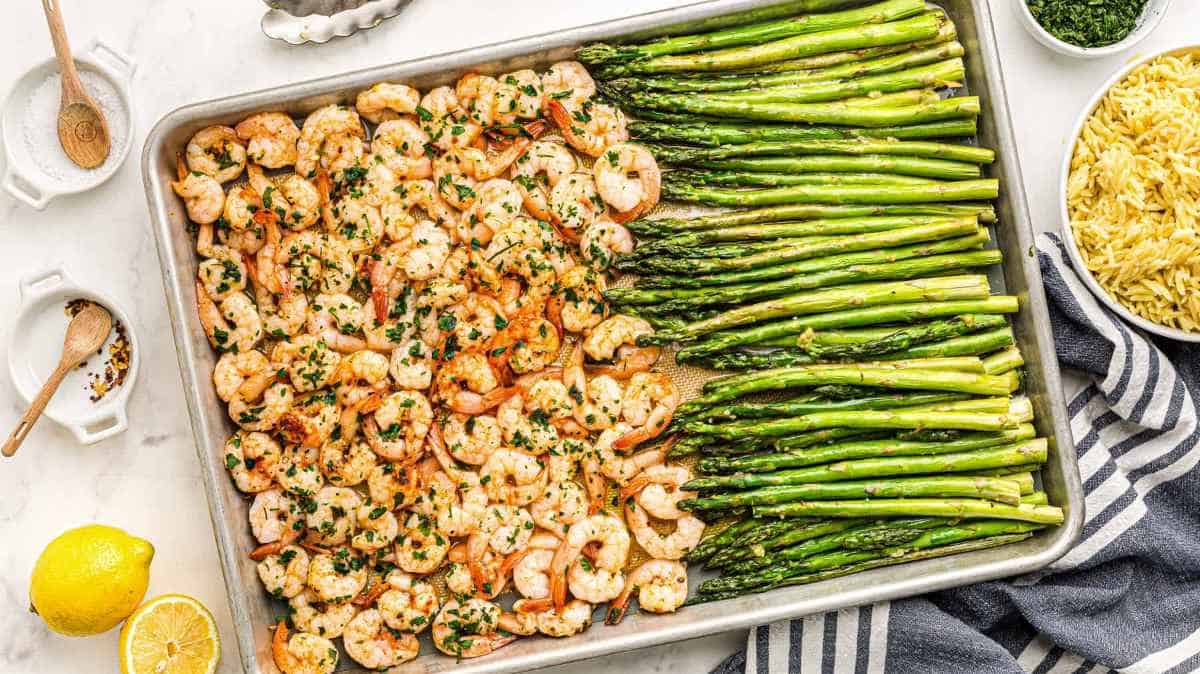 A tray of roasted shrimp and asparagus garnished with herbs, alongside lemon and pasta.