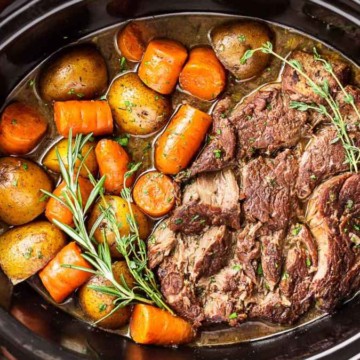 Pot roast with carrots and potatoes in a slow cooker, garnished with fresh herbs.