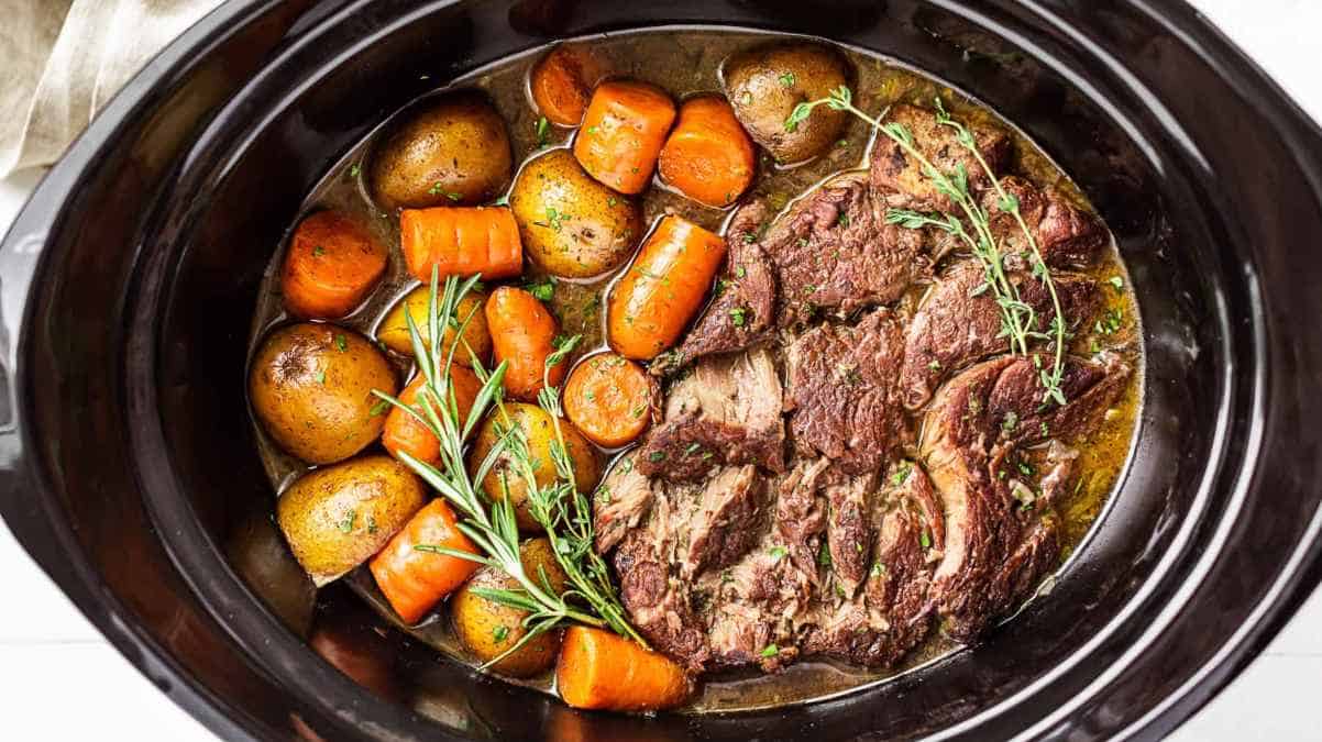 Pot roast with carrots and potatoes in a slow cooker, garnished with fresh herbs.