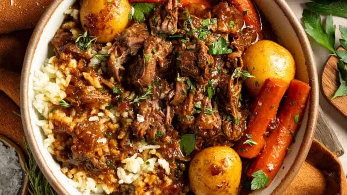 A bowl of braised beef with carrots, potatoes, and garnished with fresh herbs, served over a bed of rice.