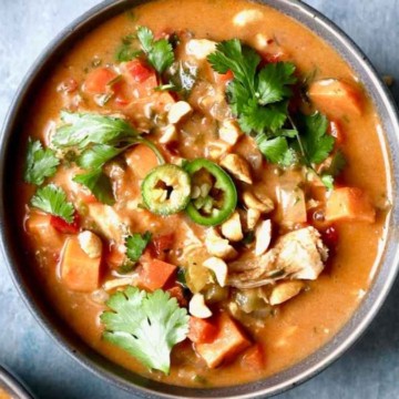 A bowl of chicken and vegetable stew garnished with cilantro and sliced jalapeños, with chopped nuts scattered around.