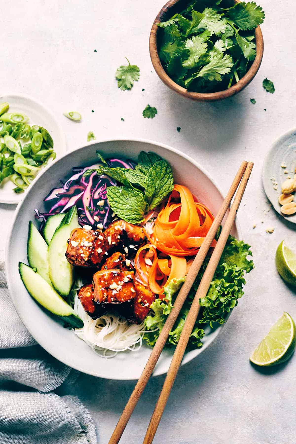 Peanut glazed tofu bowl on the counter with garnishes like cilantro, scallions and chopped nuts and limes surrounding.
