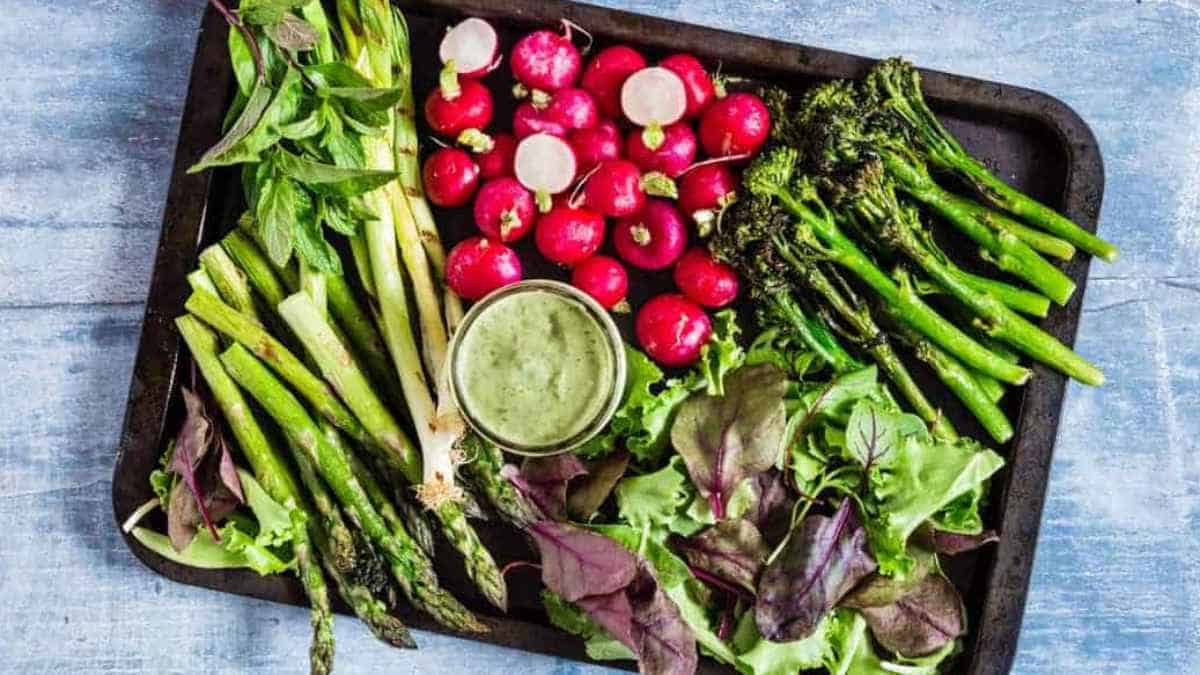 A variety of fresh vegetables with a side of green dressing served on a rustic tray.
