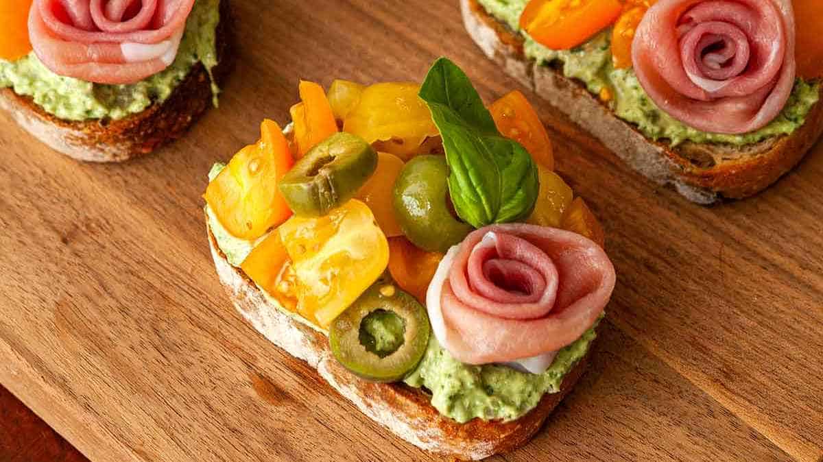 Open-faced sandwiches topped with avocado spread, assorted colorful tomatoes, and ham roses on a wooden board.