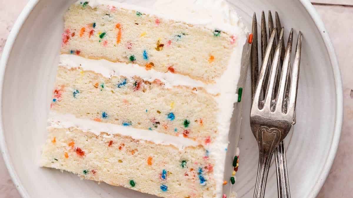 A slice of funfetti cake with white frosting on a plate next to two forks.