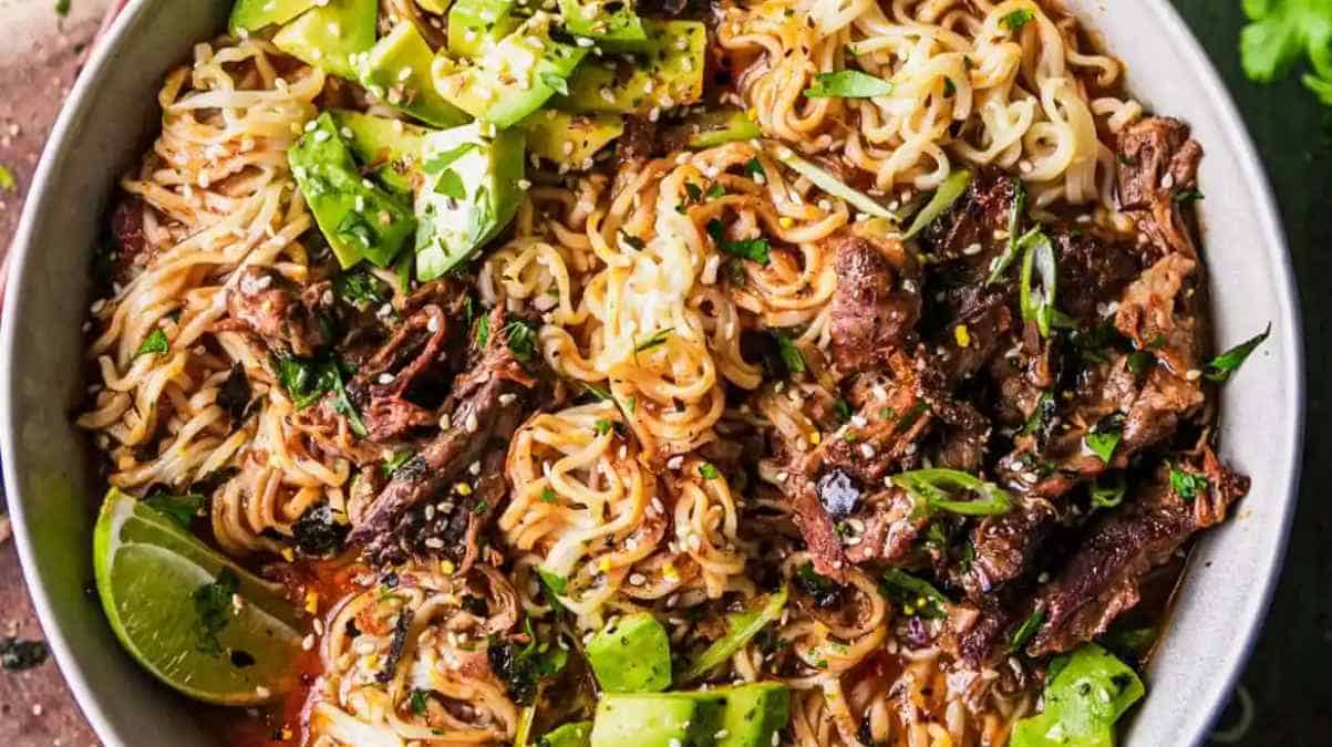 Bowl of noodles with beef, avocado, lime, and garnished with sesame seeds and herbs.