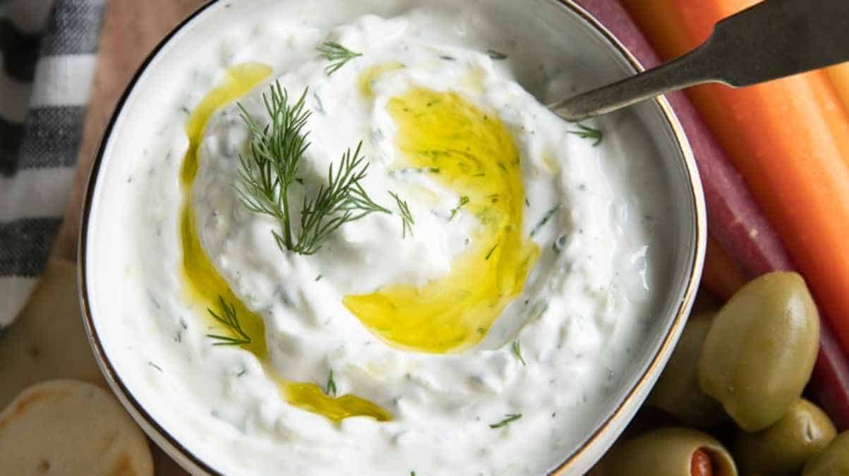A bowl of tzatziki sauce garnished with dill and olive oil, accompanied by vegetables and olives.