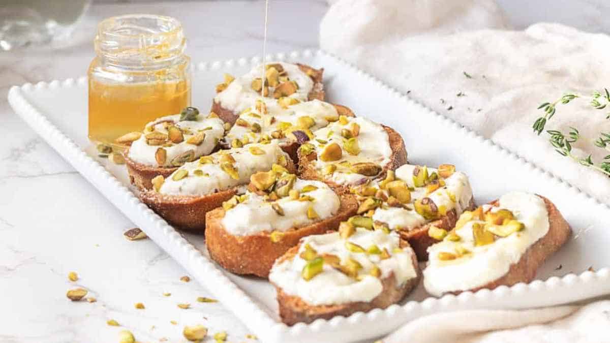 Slices of baguette topped with cream cheese and chopped pistachios, served with honey on the side.