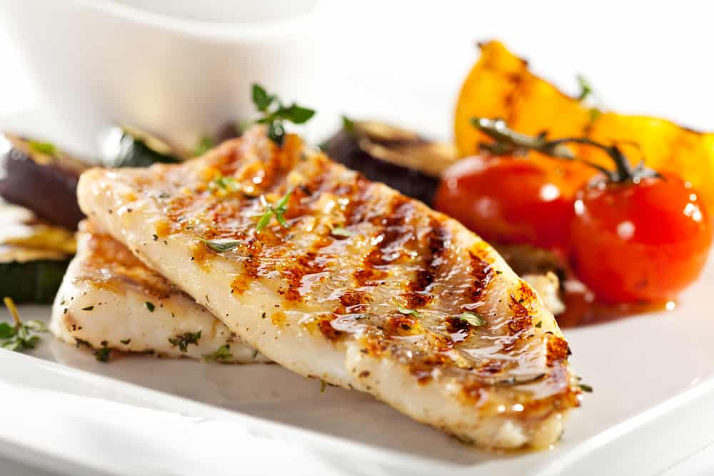 Grilled fish fillet served with zucchini and other roasted vegetables on a white plate.