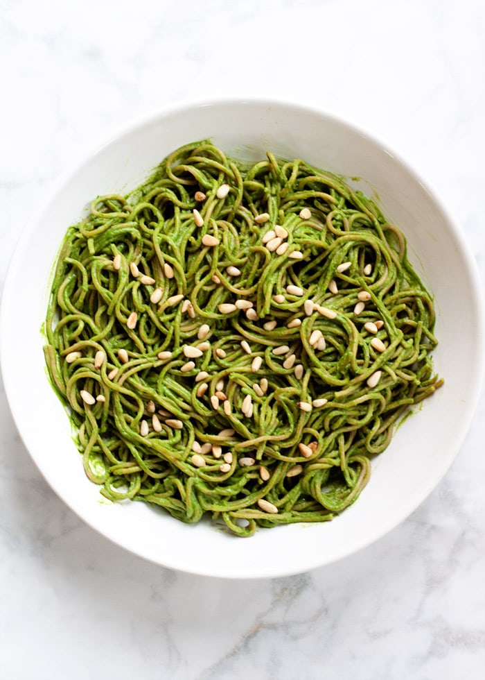 A bowl of green pasta, also known as avocado pesto pasta, in a white bowl on a marble background. Toasted pine nuts garnish the top.