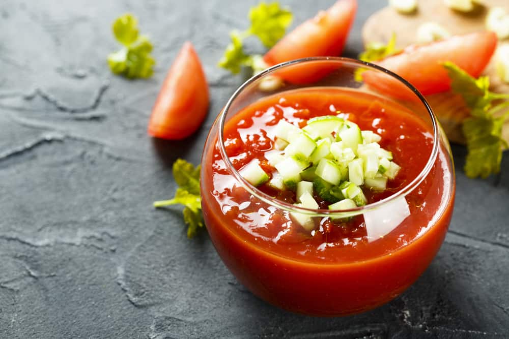 A bowl of gazpacho garnished with diced cucumber and garlic on a textured black surface with fresh ingredients in the background.