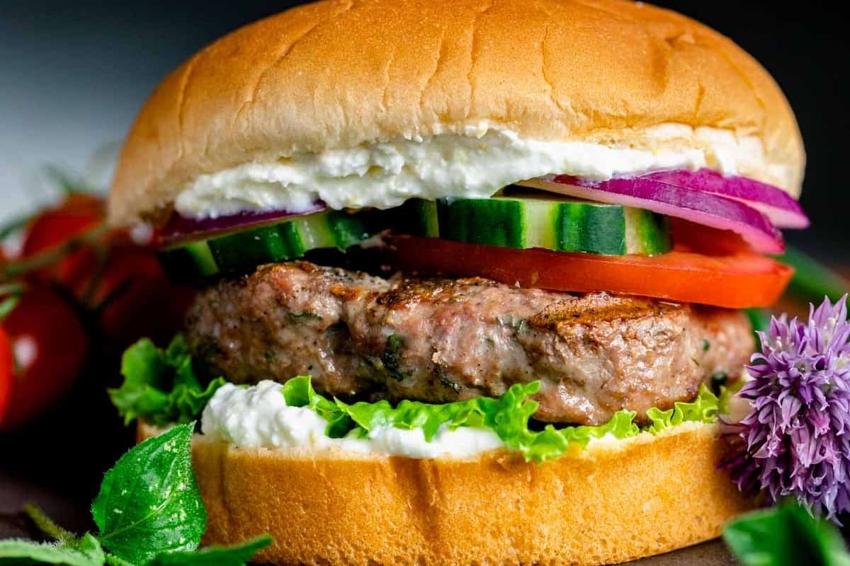 Close-up of a hamburger with a beef patty, lettuce, tomatoes, cucumbers, red onions, and a dollop of cream on a bun, accompanied by fresh vegetables on the side.