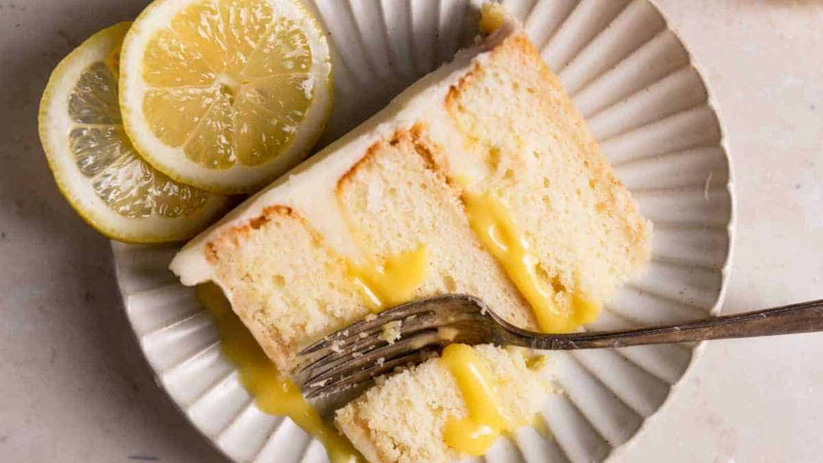 A slice of lemon cake with lemon glaze on a plate with a fork and sliced lemons.