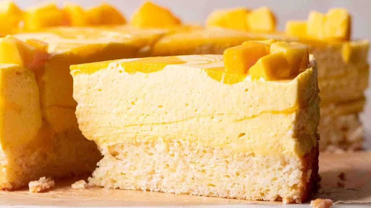 A slice of mango cheesecake garnished with mango cubes on a wooden surface.