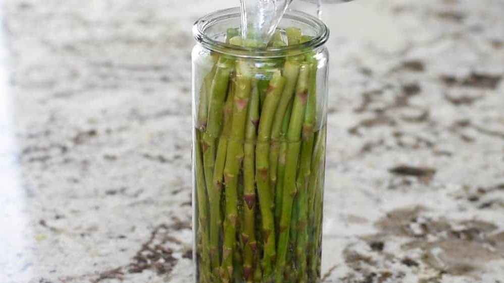 Asparagus stalks being placed into a tall glass jar with water.