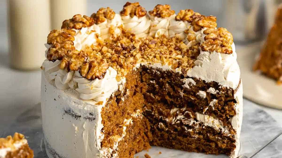 A carrot cake with cream cheese frosting, topped with walnuts, with one slice partially removed.